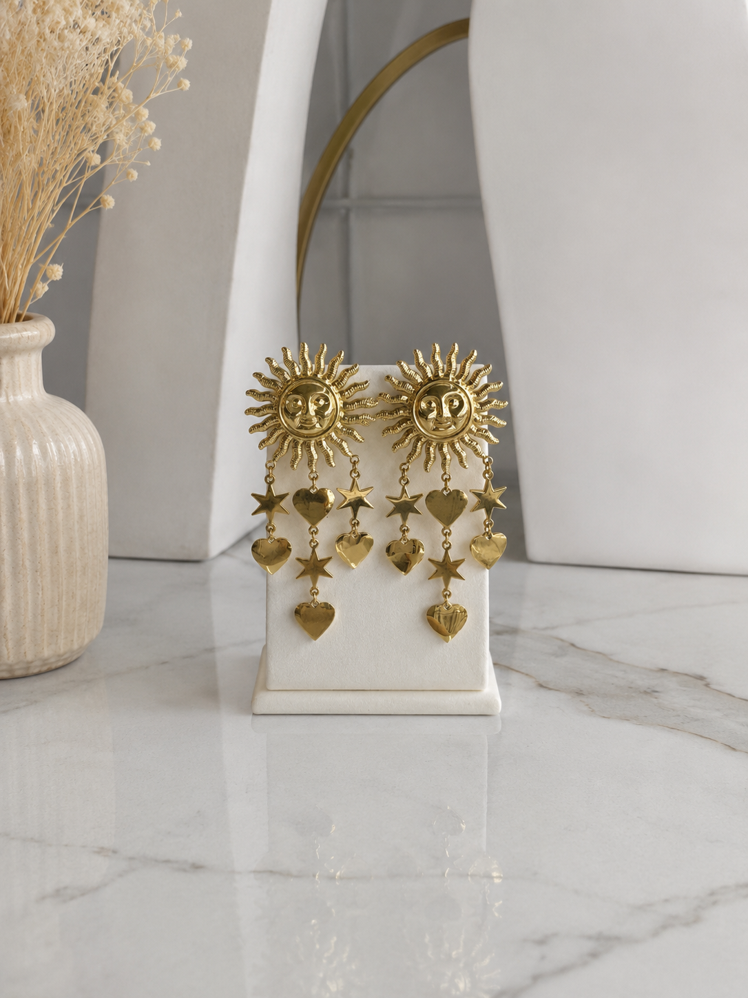 Aretes dorados grandes con medallón de sol con rostro y charms colgantes de estrellas y corazones sobre soporte de exhibición