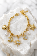 Pulsera dorada con charms celestiales de luna con cristales, estrellas, corazones y perlas sobre piedra blanca